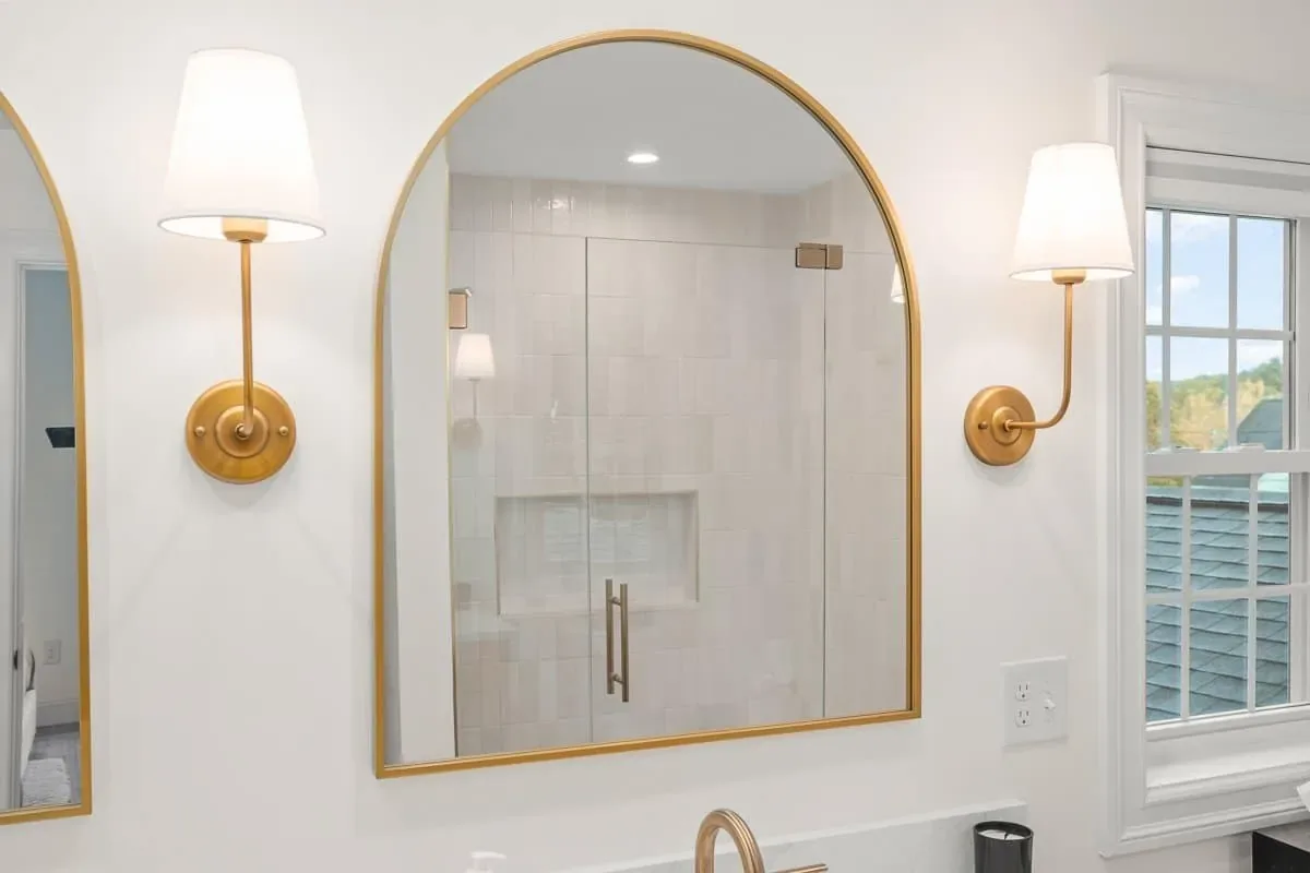 Arched brass mirror and wall sconces over the vanity, shower beyond